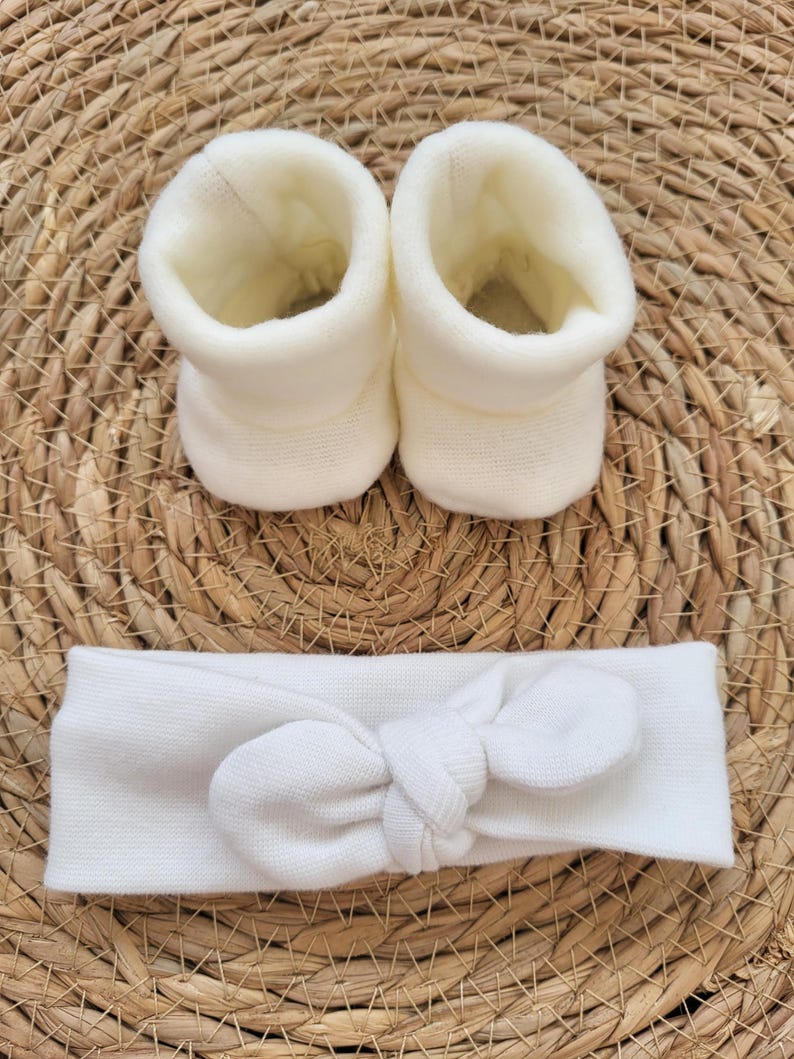 May include: A set of baby accessories, including two ivory-colored booties and a matching headband with a bow. The items are made of a soft, knitted material and are displayed on a woven, light brown surface.