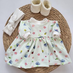 May include: A baby outfit set featuring a light green dress with a floral pattern, white booties, and a matching headband. The dress has long sleeves and a round neckline. The set is arranged on a woven mat.