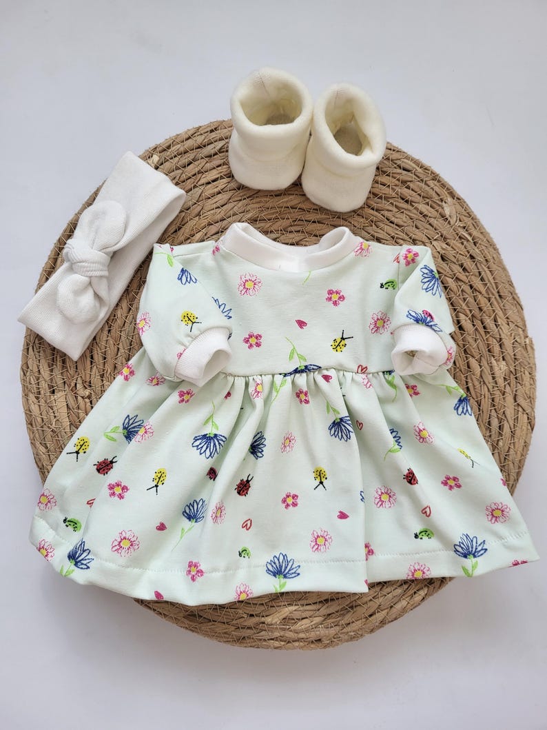 May include: A baby outfit set featuring a light green dress with a floral pattern, white booties, and a matching headband. The dress has long sleeves and a round neckline. The set is arranged on a woven mat.