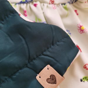 May include: Close-up of a quilted, dark teal fabric with a stitched pattern. A small, tan leather tag with a heart design is attached. The background features a white floral print fabric with pink and yellow flowers.