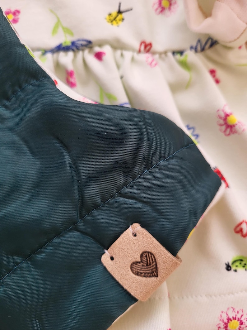 May include: Close-up of a quilted, dark teal fabric with a stitched pattern. A small, tan leather tag with a heart design is attached. The background features a white floral print fabric with pink and yellow flowers.