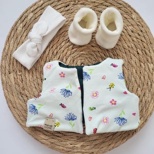 May include: A baby clothing set featuring a white headband with a knotted bow, two white booties, and a pair of floral-patterned baby shoes. The shoes have a light green background with blue, pink, and yellow flowers, and a small tag that says "Made with LOVE".