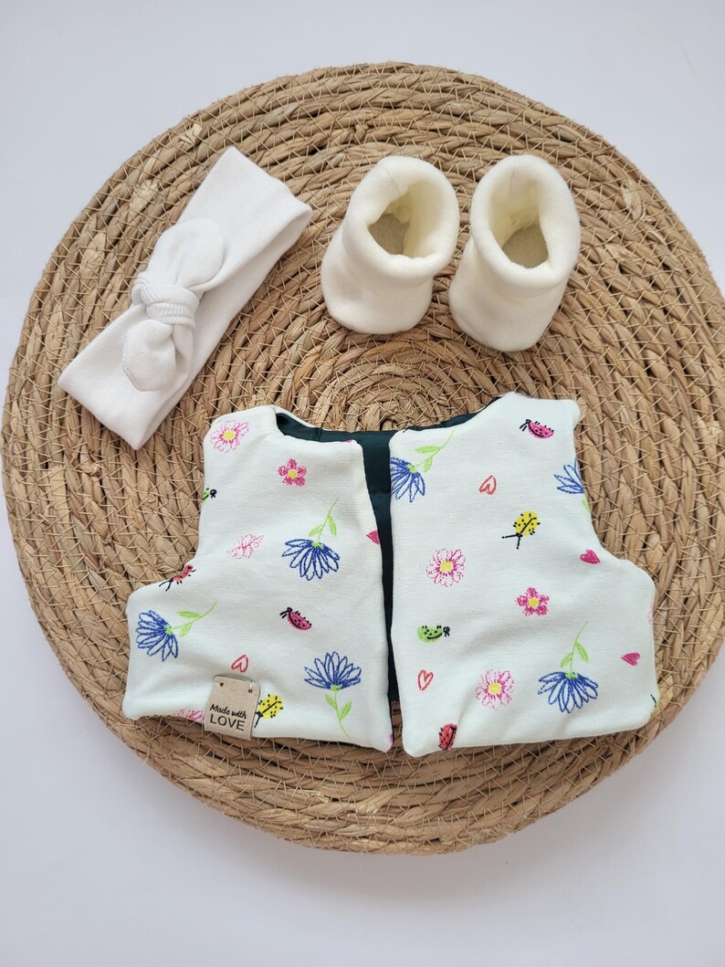 May include: A baby clothing set featuring a white headband with a knotted bow, two white booties, and a pair of floral-patterned baby shoes. The shoes have a light green background with blue, pink, and yellow flowers, and a small tag that says "Made with LOVE".