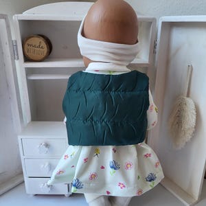 May include: A doll wearing a white turtleneck, headband, and a dark green quilted vest. The doll is also wearing a floral print dress with pink and blue flowers. The doll is standing in front of a small white cabinet.
