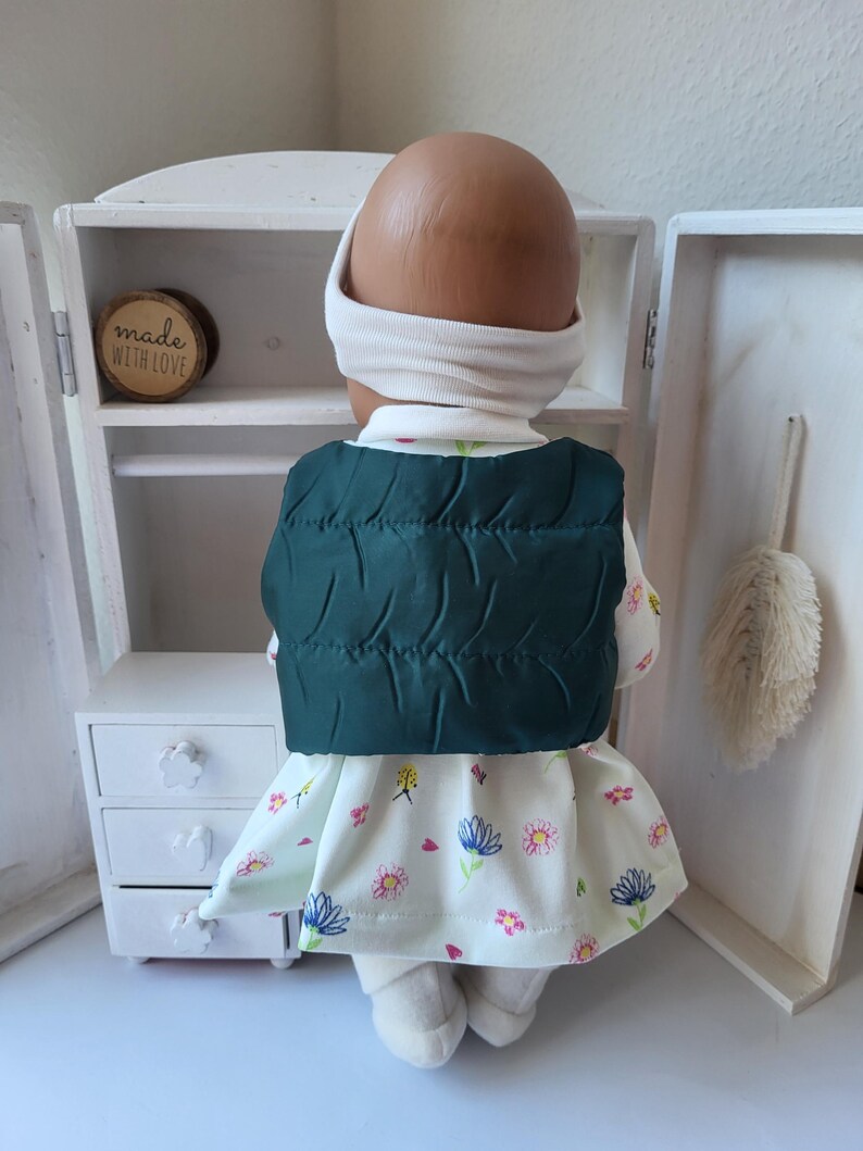 May include: A doll wearing a white turtleneck, headband, and a dark green quilted vest. The doll is also wearing a floral print dress with pink and blue flowers. The doll is standing in front of a small white cabinet.