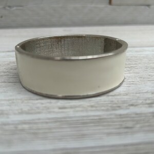 Puede incluir: Un brazalete ancho de color marfil con interior y ribete de metal plateado. El brazalete tiene un acabado liso y brillante y un cierre de bisagra. Un accesorio versátil y elegante.