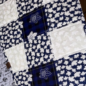 Peut inclure: Une couverture matelassée avec un motif patchwork de feuilles d'érable blanches sur fond bleu marine. La couverture comprend également des carrés à carreaux bleu marine et noir, et des carrés blancs avec des silhouettes d'ours. Le logo des Maple Leafs de Toronto est visible.