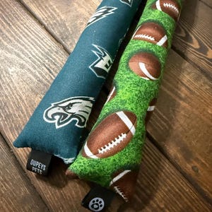 May include: Two long, cylindrical pet toys. One is teal with white Eagles logos, the other is green with brown footballs. The toys are made of fabric and have a tag that says "Dupeys Pets".