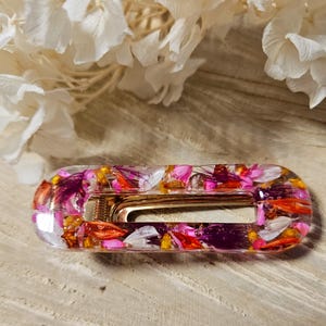 Peut inclure: Une barrette rectangulaire avec un fermoir doré, enfermée dans de la résine transparente. La résine est remplie de pétales de fleurs colorés dans les tons de rose, d'orange et de blanc. La barrette est posée sur une surface en bois.
