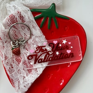 May include: A clear acrylic keychain with the text "For Your Validation" in pink and black lettering. The keychain is decorated with pink hearts and white stars. The keychain is on a red strawberry-shaped dish.
