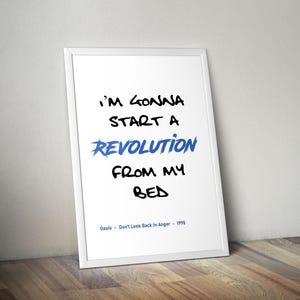 May include: A framed print with the words "I'M GONNA START A REVOLUTION FROM MY BED". The word "REVOLUTION" is in blue. The text "Oasis - Don't Look Back in Anger - 1995" is at the bottom. The frame is white.