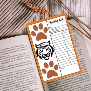 Pode incluir: Um marcador de livros com borda laranja, apresentando um rosto de tigre e pegadas, repousa sobre um livro aberto. O marcador de livros tem um layout de lista de leitura com espaços para título, data e notas. Óculos com armação preta à esquerda.