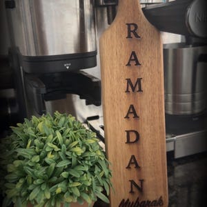 May include: Wooden Ramadan sign with the word "Ramadan Mubarak" engraved. The sign is a light brown color and has a handle. A small potted plant with green leaves is in the foreground, and a coffee machine is in the background.