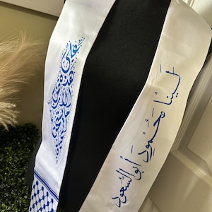 May include: White graduation sash with blue Arabic calligraphy and the year 2025 printed on it. The sash has a blue and white checkered pattern on one end.