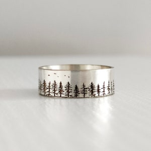 Puede incluir: Un anillo de banda de plata con un diseño de silueta de bosque. El anillo presenta una línea de árboles con pájaros volando por encima.