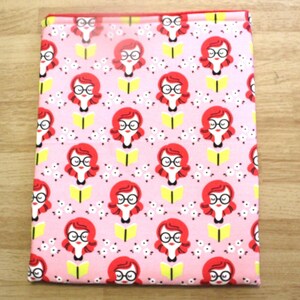 May include: A pink fabric item with a repeating pattern of a woman with red hair, glasses, and a black bow tie, reading a yellow book. Small white flowers are scattered around the figures. The item is rectangular and appears to be a book cover or a similar accessory.