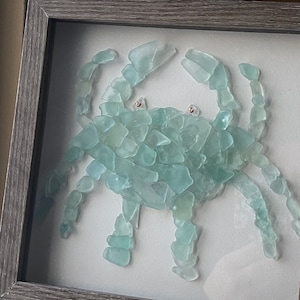 May include: A framed artwork of a crab made from blue sea glass. The crab is displayed on a white background.