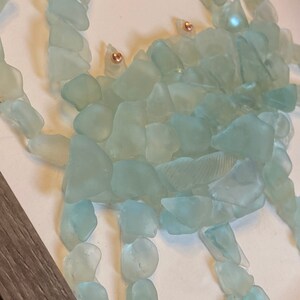May include: A sea-themed wall art piece featuring a crab made from light blue sea glass. The crab has copper-colored eyes and is set against a white background.