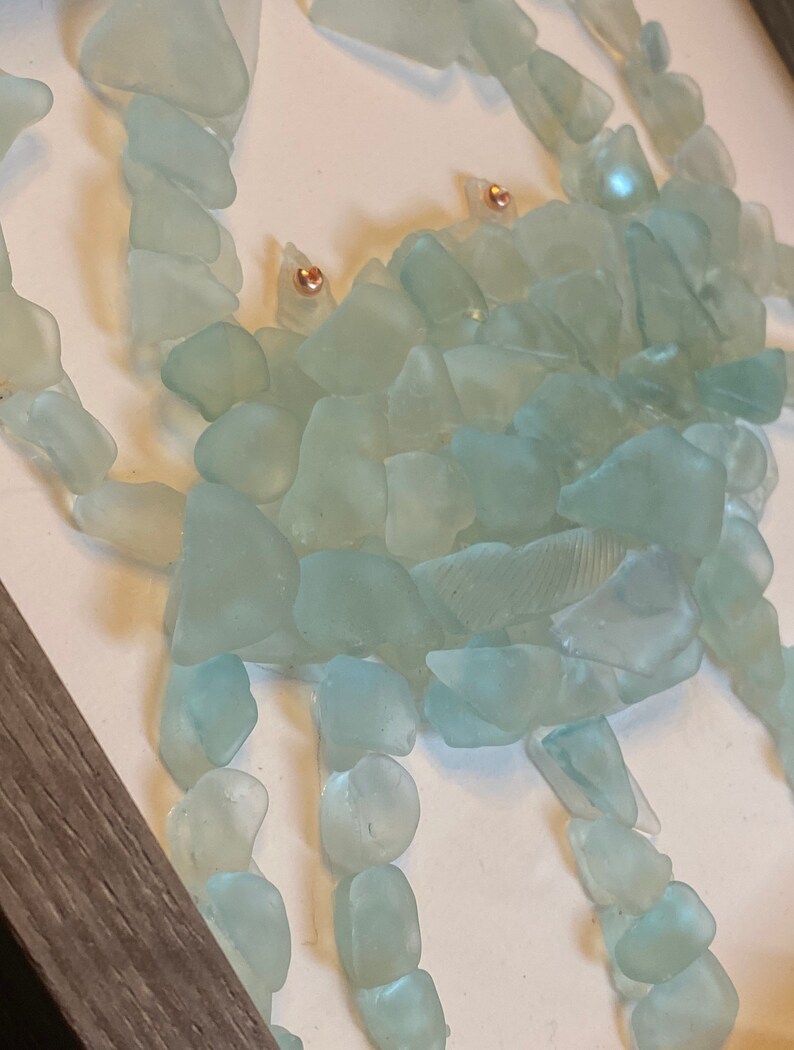 May include: A sea-themed wall art piece featuring a crab made from light blue sea glass. The crab has copper-colored eyes and is set against a white background.