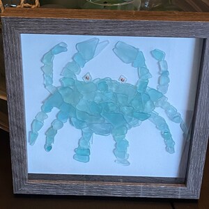 May include: A framed artwork depicting a blue crab made from sea glass. The crab is in a light blue color and is set against a white background.
