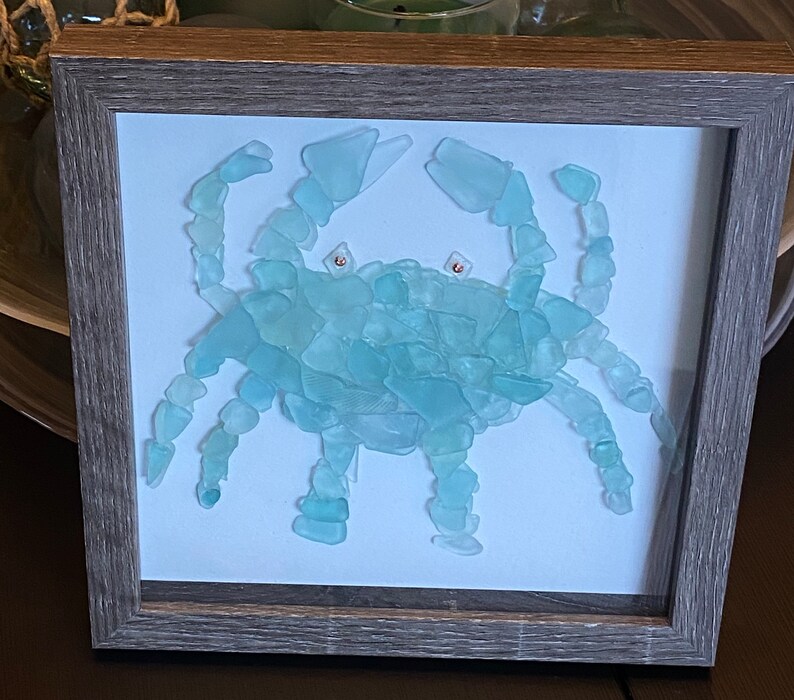 May include: A framed artwork depicting a blue crab made from sea glass. The crab is in a light blue color and is set against a white background.