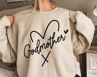 May include: A beige crewneck sweatshirt featuring black text that reads Godmother in a cursive script. Above the text is a simple black line drawing of a heart shape, and below the text is a small black letter x. The sweatshirt has long sleeves.