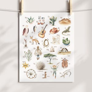 May include: Watercolor alphabet poster featuring Western-themed illustrations. Includes images of boots, a guitar, a cactus, a lizard, a sheriff's star, and a tumbleweed. The poster is hanging from a clothesline with wooden clips.