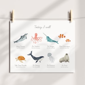 May include: A white poster with watercolor illustrations of sea creatures, including a dolphin, octopus, narwhal, and clownfish. Text reads "Today I will" with inspirational phrases like "Stay Curious" and "Be Creative". The poster is held by wooden clips.