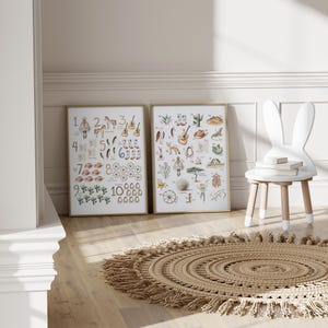 May include: Two framed art prints with gold frames, featuring watercolor illustrations of numbers and western-themed objects. A white bunny-shaped chair and a round, fringed rug are also in the room. The prints are on a light wood floor.