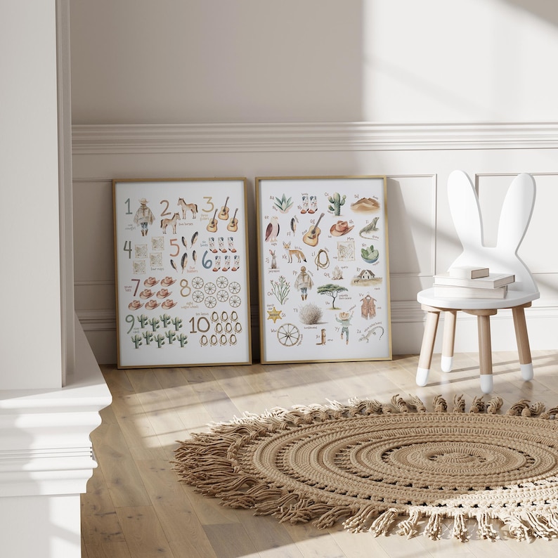 May include: Two framed art prints with gold frames, featuring watercolor illustrations of numbers and western-themed objects. A white bunny-shaped chair and a round, fringed rug are also in the room. The prints are on a light wood floor.