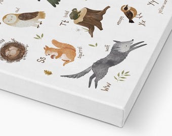 May include: A canvas print featuring an illustrated alphabet with woodland animals and plants. The letters are paired with images of an owl, squirrel, wolf, and other creatures. The artwork has a watercolor style with a white background.