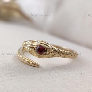 Puede incluir: Anillo de serpiente de oro con un ojo de piedra preciosa roja. La serpiente está enroscada y la cola está enroscada. El anillo está hecho de oro y tiene un diseño detallado.