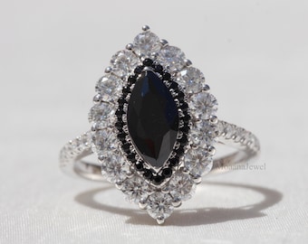 Pode incluir: Um anel de prata com uma pedra preta em forma de marquesa, rodeada por pedras brancas e pretas. A banda do anel é cravejada com pequenas pedras transparentes. O anel é apresentado sobre um fundo branco.