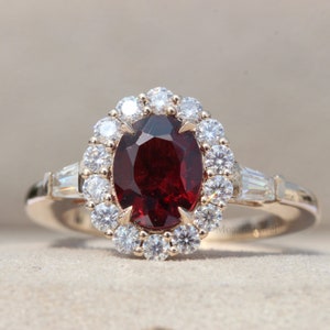 May include: A gold ring with a large oval red gemstone surrounded by a halo of smaller diamonds. The ring has two baguette-shaped diamonds on each side of the center stone.