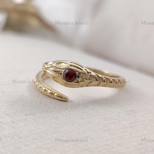 Könnte beinhalten: Ein goldener Ring in Schlangenform mit einem strukturierten, schuppenartigen Muster entlang der Ringschiene und einem einzelnen, runden, tiefroten Edelstein, der in den Kopf der Schlange eingefasst ist. Der Ring hat ein offenes Design.