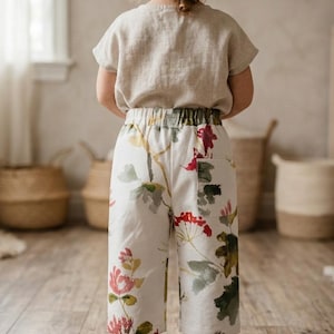 Może przedstawiać: Dziecko w beżowej lnianej górze i białych spodniach w kwiaty w kolorze czerwonym, zielonym i różowym. Dziecko ma również na sobie żółte mokasyny. Strój jest odpowiedni do noszenia na co dzień.