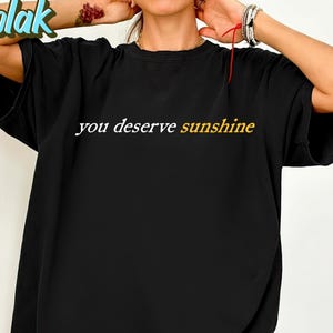 May include: Black t-shirt with the text "you deserve sunshine" in white and yellow. The shirt has short sleeves and a relaxed fit. The brand name "Nodolak" is visible in the upper left corner.