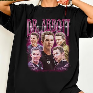 May include: Black t-shirt featuring a graphic of a man with the text "DR. ABBOTT" in a pink and white font. The graphic includes multiple images of the same man. The shirt is a casual style.