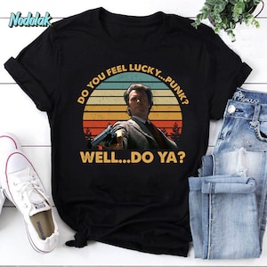 Peut inclure: Un t-shirt noir avec un graphique vintage de Clint Eastwood en tant que Dirty Harry. Le graphique présente un arrière-plan de coucher de soleil avec le texte "Do you feel lucky...punk? Well...do ya?"