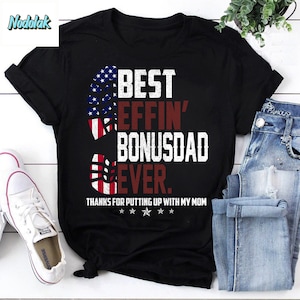 May include: Black t-shirt with a red, white, and blue American flag design. The text "BEST EFFIN' BONUSDAD EVER." is printed on the shirt. Below the text is "THANKS FOR PUTTING UP WITH MY MOM" and five stars.