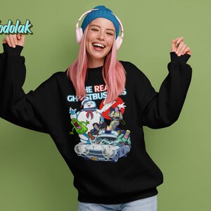 K&ouml;nnte beinhalten: Schwarzer Pullover mit einem Aufdruck der Ghostbusters-Charaktere, darunter Slimer, die im Ecto-1 fahren. Der Text "The Real Ghostbusters" ist in Neonblau &uuml;ber den Figuren gedruckt.