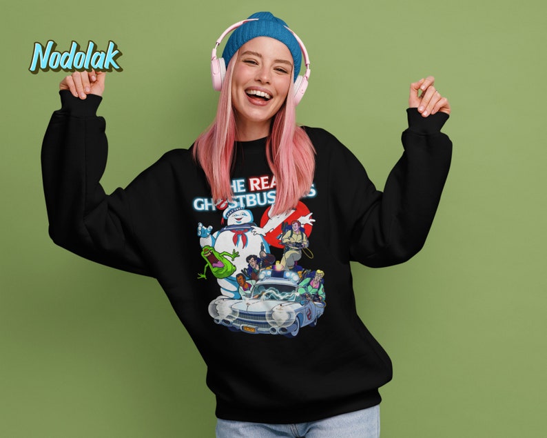 K&ouml;nnte beinhalten: Schwarzer Pullover mit einem Aufdruck der Ghostbusters-Charaktere, darunter Slimer, die im Ecto-1 fahren. Der Text "The Real Ghostbusters" ist in Neonblau &uuml;ber den Figuren gedruckt.