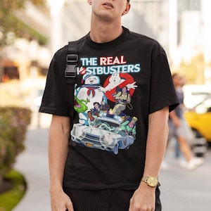 K&ouml;nnte beinhalten: Ein schwarzes T-Shirt mit einem Grafikdruck der Ghostbusters-Charaktere, darunter Slimer, in ihrem legend&auml;ren Auto. Der Text "THE REAL GHOSTBUSTERS" ist in Rot und Blau auf dem Shirt gedruckt.