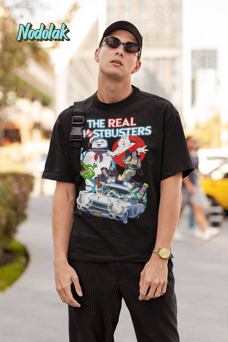 K&ouml;nnte beinhalten: Ein schwarzes T-Shirt mit einem Grafikdruck der Ghostbusters-Charaktere, darunter Slimer, in ihrem legend&auml;ren Auto. Der Text "THE REAL GHOSTBUSTERS" ist in Rot und Blau auf dem Shirt gedruckt.