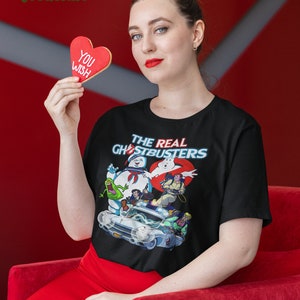 K&ouml;nnte beinhalten: Ein schwarzes T-Shirt mit einem Grafikdruck der Ghostbusters-Charaktere und ihres Autos. Der Text "The Real Ghostbusters" ist auf dem Shirt gedruckt. Eine Person h&auml;lt einen roten herzf&ouml;rmigen Keks, auf dem "You Wish" steht.