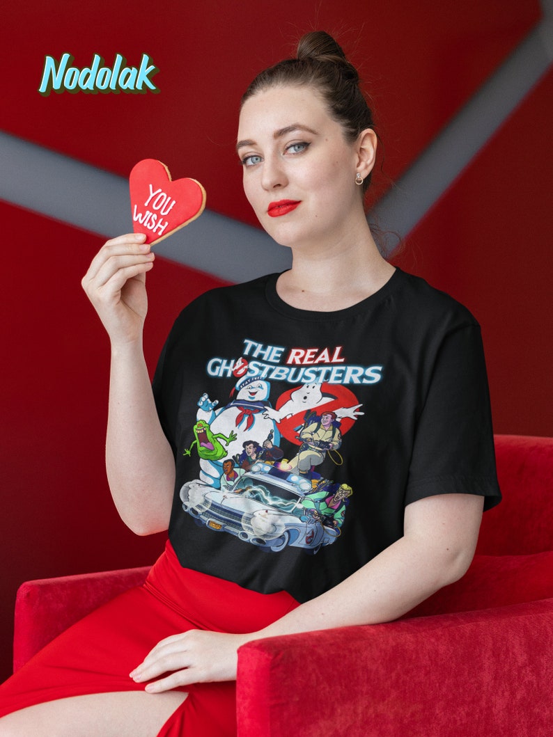 K&ouml;nnte beinhalten: Ein schwarzes T-Shirt mit einem Grafikdruck der Ghostbusters-Charaktere und ihres Autos. Der Text "The Real Ghostbusters" ist auf dem Shirt gedruckt. Eine Person h&auml;lt einen roten herzf&ouml;rmigen Keks, auf dem "You Wish" steht.