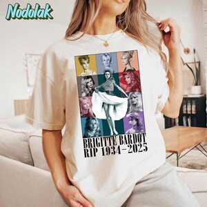 K&ouml;nnte beinhalten: Cremefarbenes T-Shirt mit einem farbenfrohen Grafik von Brigitte Bardot. Die Grafik enth&auml;lt mehrere Bilder der Schauspielerin und den Text "BRIGITTE BARDOT RIP 1934-2025". Das Shirt hat kurze &Auml;rmel und einen Rundhalsausschnitt.