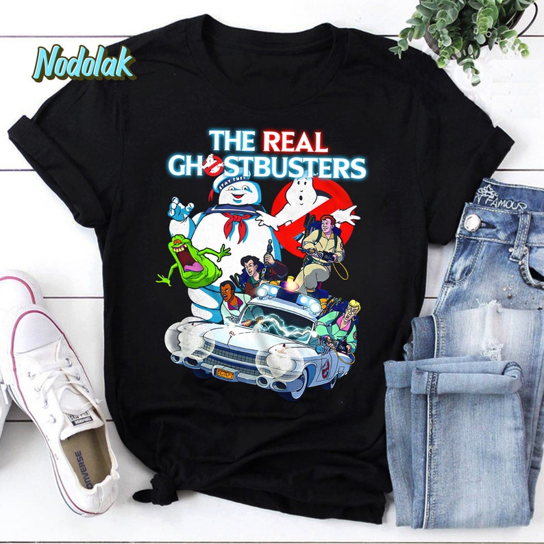 K&ouml;nnte beinhalten: Schwarzes T-Shirt mit einem Grafikdruck der Ghostbusters-Charaktere aus der Zeichentrickserie. Der Text "THE REAL GHOSTBUSTERS" ist in Wei&szlig; auf dem Shirt gedruckt. Die Figuren stehen vor dem Ghostbusters-Auto, dem Ecto-1.