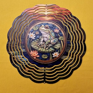 May include: A metal wind spinner with a circular design featuring a green frog sitting on a lily pad surrounded by water lilies. The background is a sunset with orange, yellow, and red hues. The wind spinner has multiple layers of metal that spin in the wind.