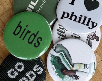 May include: A collection of six circular pin-back buttons featuring various designs. One is dark green with white text reading 'PHILLY the tush push'. Another is white with 'I heart philly' text. A bright green button says 'birds'. A black button says 'go birds'. Two white buttons feature football imagery.